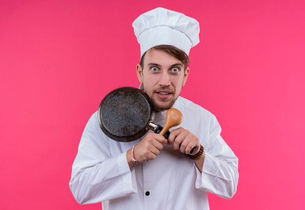 Een grappige jonge, bebaarde chef-kok in wit uniform toont x-bord met koekenpan en houten lepel terwijl hij op een roze muur kijkt