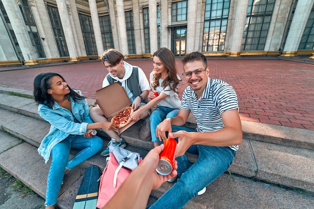 Een groep studenten zit op de trappen buiten de campus en eet pizza en frisdrank. een groep vrienden is aan het ontspannen en aan het kletsen.