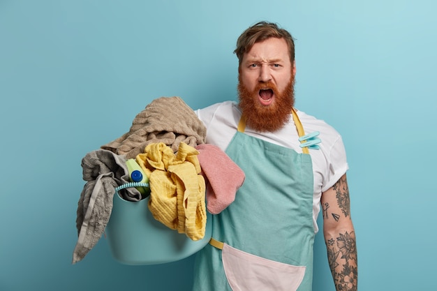 Een hardwerkende roodharige man met een baard doet huishoudelijke klusjes, is bezig met wassen, houdt een mand met wasgoed vast, draagt een schort, wasknijpers, roept luid uit, wordt overvol met het huishouden. Huishoudelijk concept.