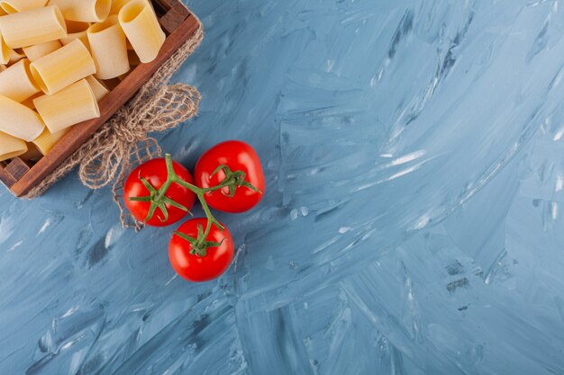 Een houten doos van droge rauwe pasta met verse rode tomaten op een marmeren tafel.