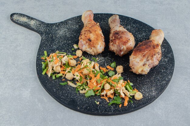 Een houten plank met gebakken kippenpoten en groentesalade.