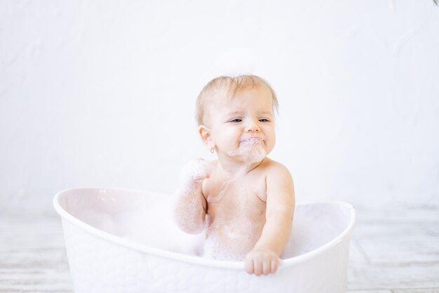 Een huilend klein babymeisje met schuim in haar mond en voor haar ogen baadt in een badkuip met schuim en zeepbellen in een lichte badkamer, het concept van kinderhygiëne