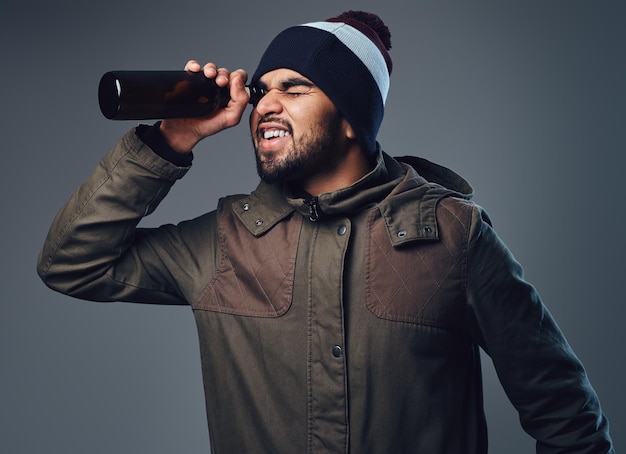 Een Indiase man met een baard, gekleed in een warme jas en een hoed die door een bierfles kijkt.