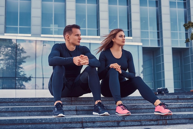 Een jong fitnesspaar in sportkleding, zit op trappen in de moderne stad tegen een wolkenkrabber. Gezond levensstijlconcept.