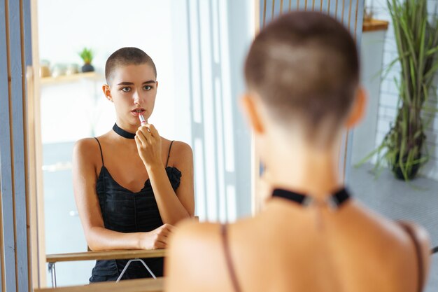 Een jong mooi meisje met kort haar schildert haar lippen, kijkend naar de weerspiegeling in de spiegel in zwarte kleding