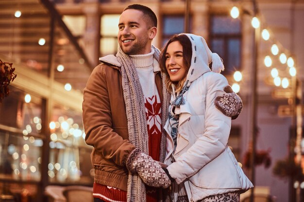 Een jong romantisch stel dat warme kleren draagt en buiten in de avondstraat knuffelt met Kerstmis