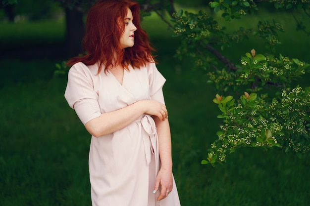 Een jong roodharig meisje in een roze kleding die zich in een de zomer groen park dichtbij bomen bevindt