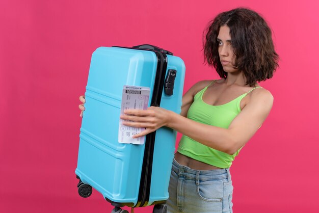 Een jonge aantrekkelijke vrouw met kort haar die groen gewasbovenkant dragen die blauwe koffer met vliegtuigtickets toont