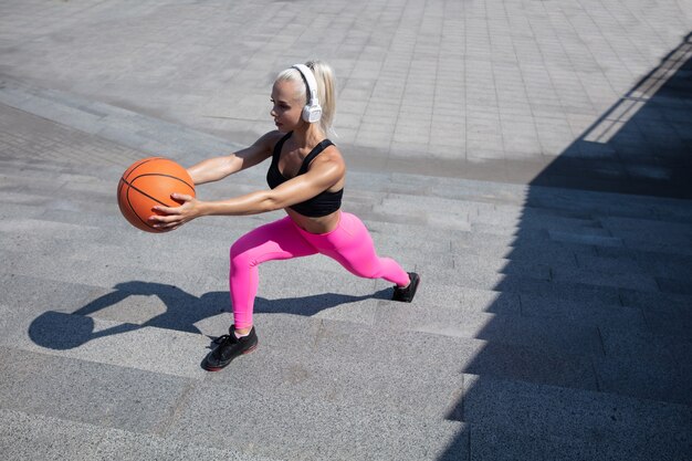 Een jonge atletische vrouw in overhemd en witte hoofdtelefoons die aan de muziek in de straat buiten werken uitwerken. Lunges doen met de bal. Concept van een gezonde levensstijl, sport, activiteit, gewichtsverlies.