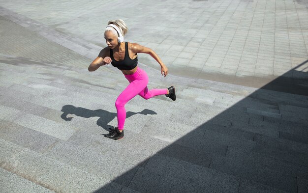 Een jonge atletische vrouw in overhemd en witte hoofdtelefoons die aan de muziek in de straat buiten werken uitwerken