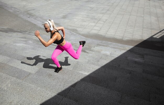Een jonge atletische vrouw in overhemd en witte hoofdtelefoons die aan de muziek in de straat buiten werken uitwerken.