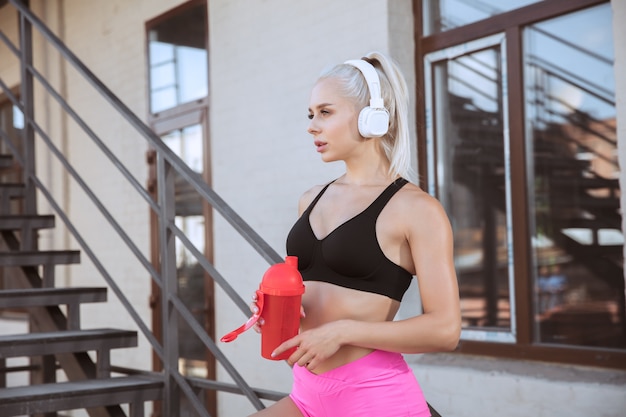 Een jonge atletische vrouw in witte koptelefoon uit te werken luisteren naar de muziek op een trap buitenshuis. Drinkwater uit de sportfles. Concept van een gezonde levensstijl, sport, activiteit, gewichtsverlies.
