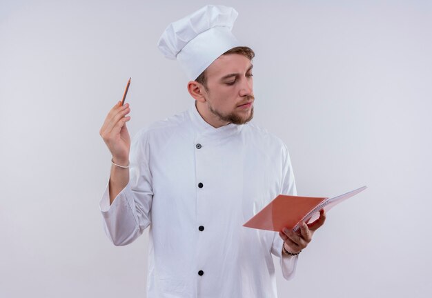Een jonge, bebaarde chef-kokmens die wit eenvormig fornuis en hoed draagt die notitieboekje bekijkt en aantekeningen maakt op een witte muur