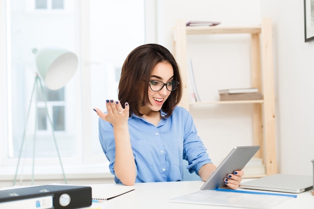 Een jonge brunette meisje in een blauw shirt zit aan de tafel op kantoor. Ze werkt met tablet. Ze kijkt aangenaam verrast.