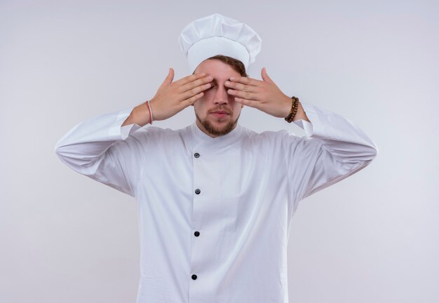 Een jonge, gebaarde chef-kokmens die witte eenvormige fornuis en hoed draagt die zijn ogen behandelt met handen op een witte muur