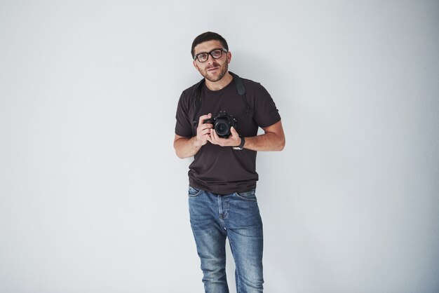 Een jonge hipster man in oculairs houdt een DSLR camera in handen staande tegen een witte muur