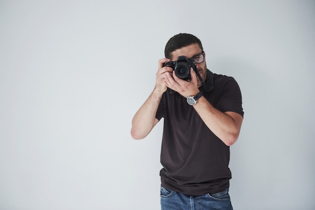 Een jonge hipster man in oculairs houdt een DSLR camera in handen staande tegen een witte muur