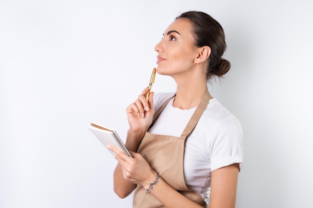 Een jonge huisvrouw in een schort op een witte achtergrond met een notitieboekje in haar handen kiest recepten voor het avondeten, bedenkt hoe ze haar man kan plezieren, bedenkt ideeën voor gerechten