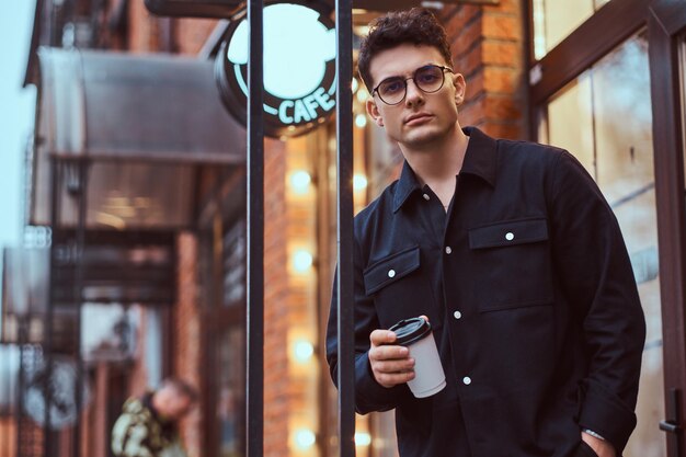 Een jonge knappe man die een afhaalkoffie vasthoudt terwijl hij buiten in de buurt van een café staat.