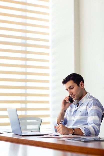 Een jonge knappe man van het vooraanzicht in gestreept shirt werken in de vergaderzaal met behulp van zijn zilveren laptop kijken door documenten praten aan de telefoon tijdens het bouwen van dagactiviteiten