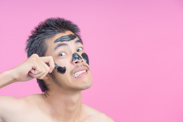 Een jonge man die zijn handen gebruikt om zwarte cosmetica op zijn gezicht te verwijderen met pijn aan een roze.