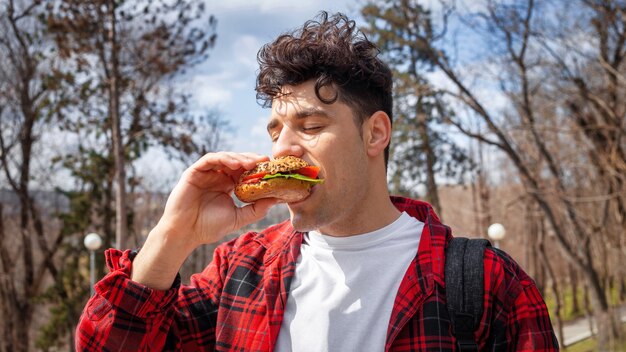 Een jonge man in een rood shirt die een hamburger eet in een park, kale bomen