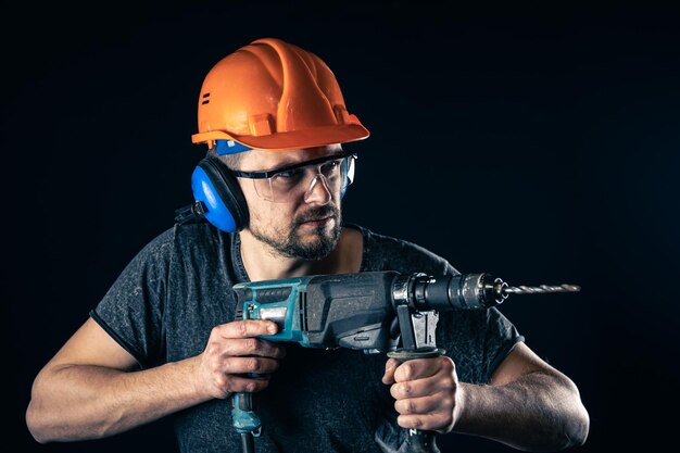 Een jonge man in overall beschermende helm harde hoed houdt elektrische boor vast