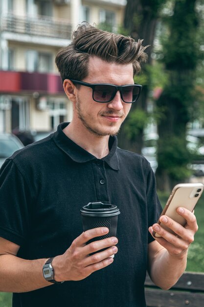 Een jonge man in zonnebril met smartphone en koffie in de stad