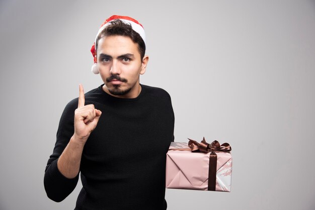 Een jonge man met een kerstmuts toont een vinger en houdt een geschenk vast.