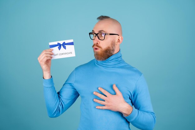 Een jonge man met een rode baard in een coltrui en een bril op een blauwe achtergrond heeft een cadeaubon, glimlacht vrolijk, parmantig humeur