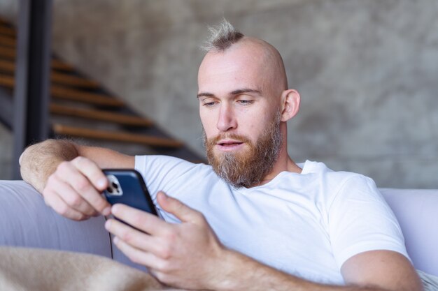 Een jonge man thuis op de bank komt naar buiten met een gezellige warme deken, houdt een mobiele telefoon vast, leest het nieuws, kijkt naar een video