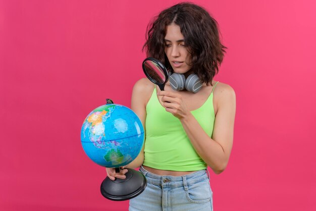Een jonge mooie vrouw met kort haar in een groene crop top in koptelefoon kijken wereldbol met vergrootglas
