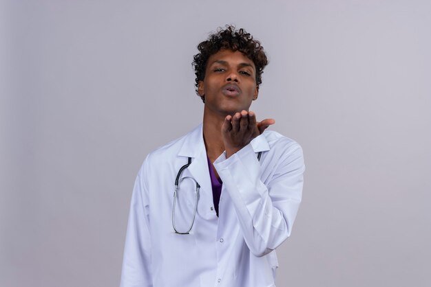 Een jonge opgewonden knappe donkere mannelijke arts met krullend haar die een witte jas draagt met een stethoscoop die een gebaar maakt van het sturen van een kus met zijn hand