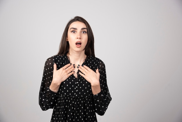 Een jonge vrouw in jurk wijst met twee handen naar zichzelf.