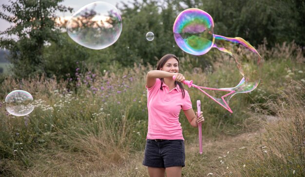 Een jonge vrouw lanceert grote gekleurde zeepbellen tussen het gras in de natuur.