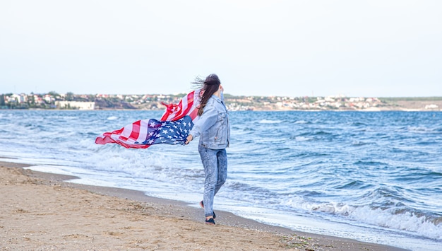 Een jonge vrouw met een Amerikaanse vlag rent langs de zee. Het concept van patriottisme en onafhankelijkheidsdagvieringen.