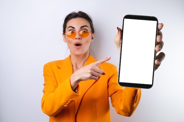 Een jonge vrouw met een trendy stijlvolle bril en een feloranje oversized jas op een witte achtergrond houdt een telefoon vast met een leeg wit scherm