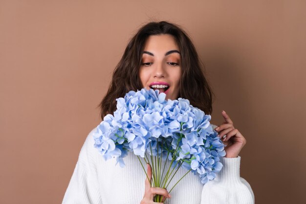 Een jonge vrouw met golvend volumineus haar op een beige achtergrond met felroze lippenstift lipgloss in een witte trui houdt een boeket blauwe bloemen vast