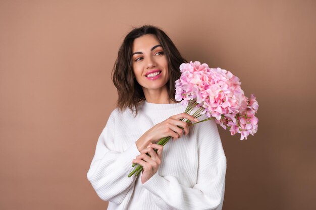 Een jonge vrouw met golvend volumineus haar op een beige achtergrond met felroze lippenstift lipgloss in een witte trui houdt een boeket roze bloemen vast