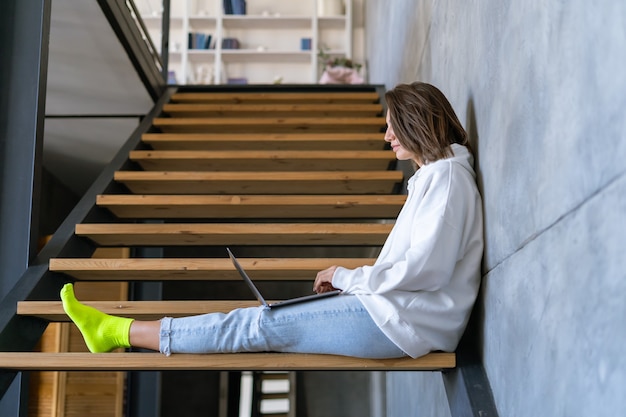 Een jonge vrouw thuis in een witte hoodie en spijkerbroek zit op de trap met een laptop op haar knieën