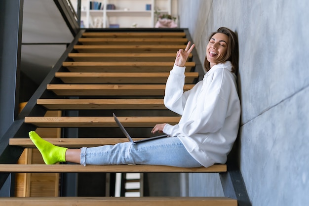 Een jonge vrouw thuis in een witte hoodie en spijkerbroek zit op de trap met een laptop op haar knieën