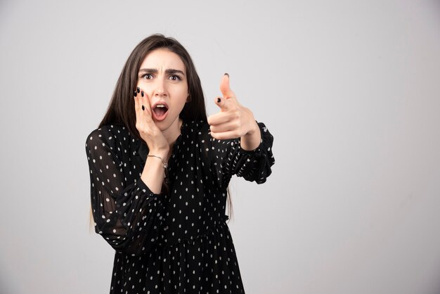 Een jonge vrouw wijst naar de camera op een grijze muur.