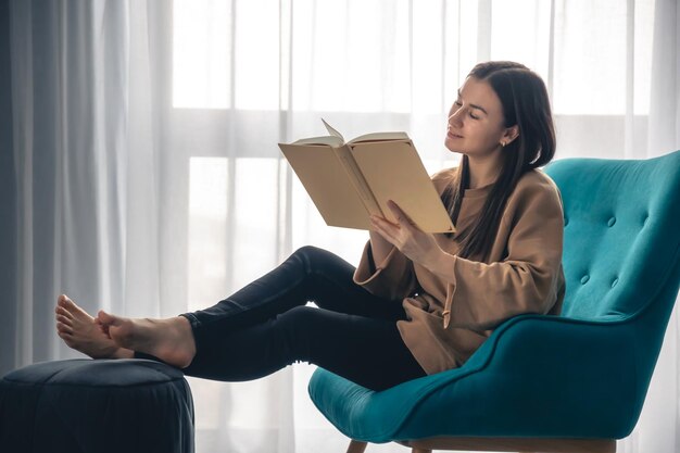 Een jonge vrouw zit een boek te lezen terwijl ze op een leunstoel bij het raam zit