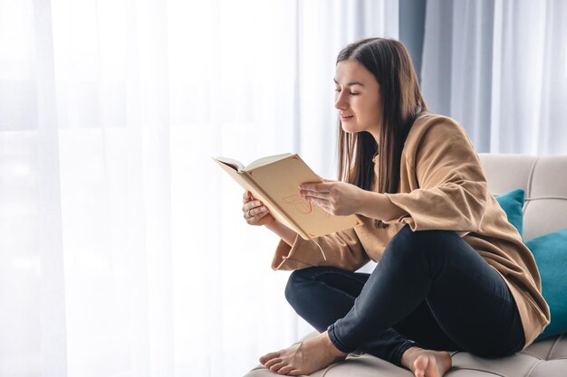 Een jonge vrouw zit een boek te lezen terwijl ze op een leunstoel bij het raam zit