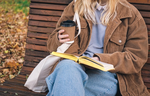 Gratis foto een jonge vrouw zit op een bankje in een herfstpark, drinkt koffie en leest een boek