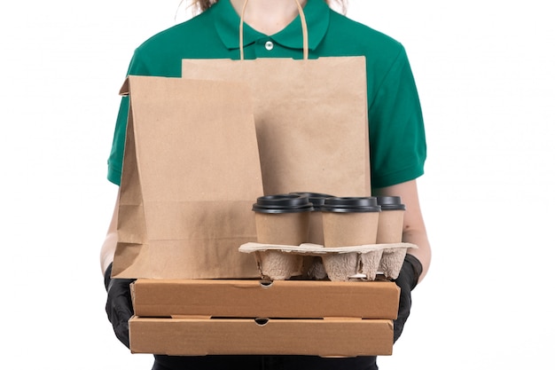 Een jonge vrouwelijke koerier van vooraanzicht in groene uniforme zwarte handschoenen en zwart masker die de pakketten van de voedsellevering het bezorgen houden