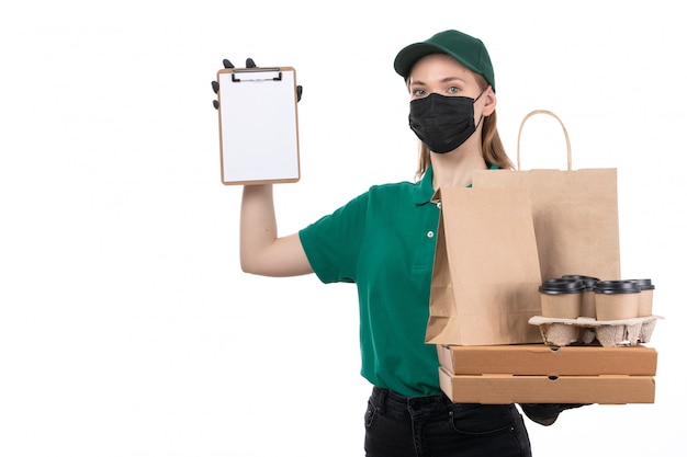 Een jonge vrouwelijke koerier van vooraanzicht in groene uniforme zwarte handschoenen en zwart masker die de pakketten van de voedsellevering het bezorgen houden