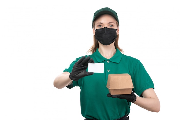 Een jonge vrouwelijke koerier vooraanzicht in groene uniforme zwarte handschoenen en zwart masker met voedsel levering pakket en witte kaart