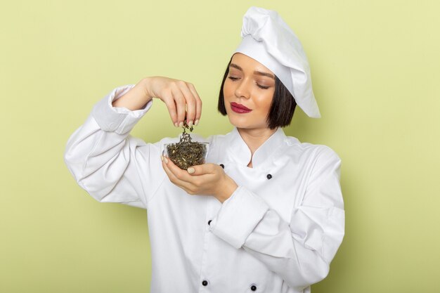 Een jonge vrouwelijke kok van vooraanzicht in wit kokkostuum en GLB-holdingskop met gedroogde thee op de groene muur
