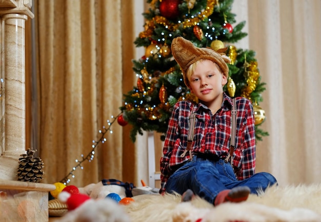 Een jongen met een grappige hertenhoed die zich voordeed op de achtergrond van de kerstboom.